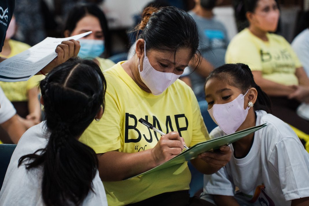 A mother in a yellow shirt signs an attendance sheet on a clipboard while children sit beside her.